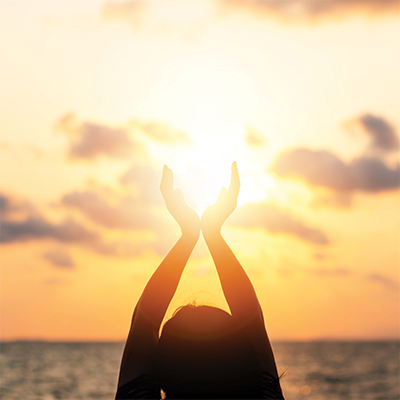 Silhoutte holding hands up to brightly lit sunrise.